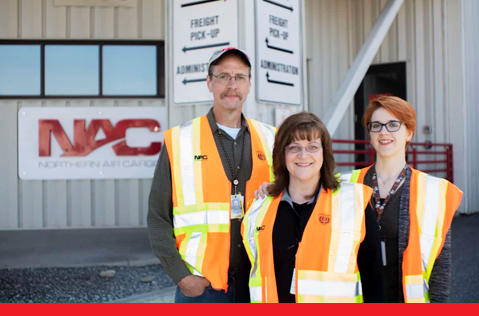 A photo of three Northern Air Cargo employees standing in front of the NAC building.